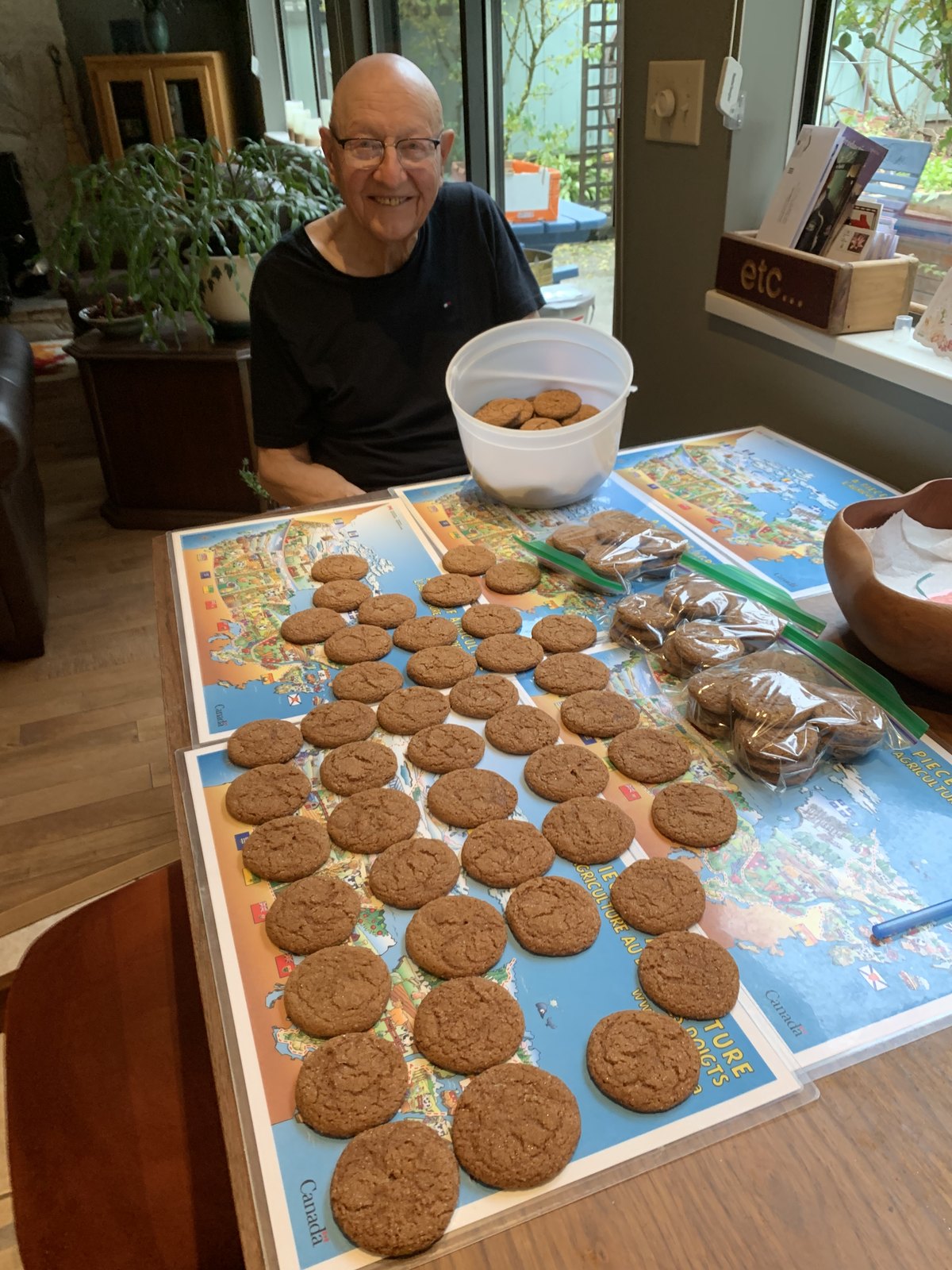 Walter, smiling behind a kitchen table covered in dozens of fresh ginger bend cookies laid out on Canada-map placemats, with an ice cream pail and sandwich bags at the ready