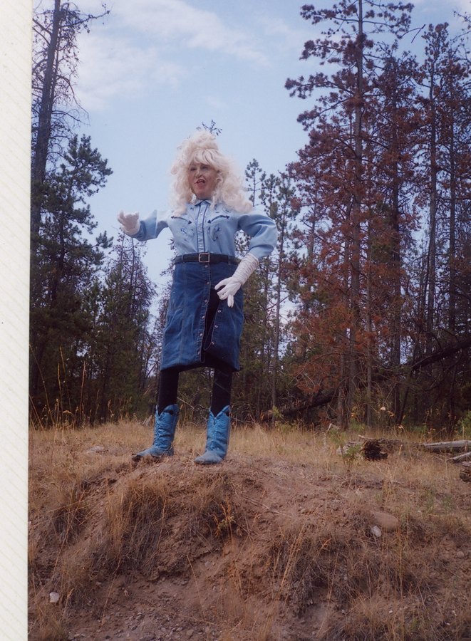 Lillooet Lill up close in her blond wig, denim western wear, long white gloves and blue cowboy boots, posed on the dry hillside