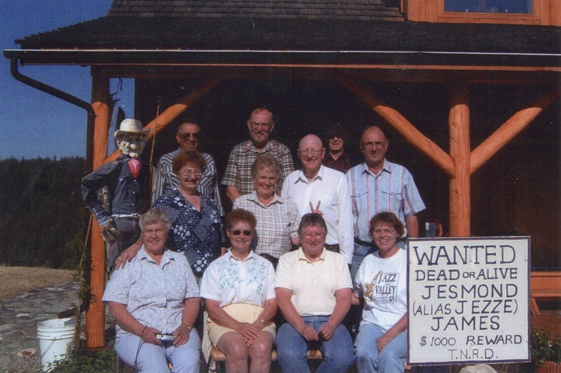 A family reunion group photo on the cabin porch — and standing at the far left among the relatives, a straw-stuffed figure in a cowboy hat, with the Jesmond James wanted sign at right