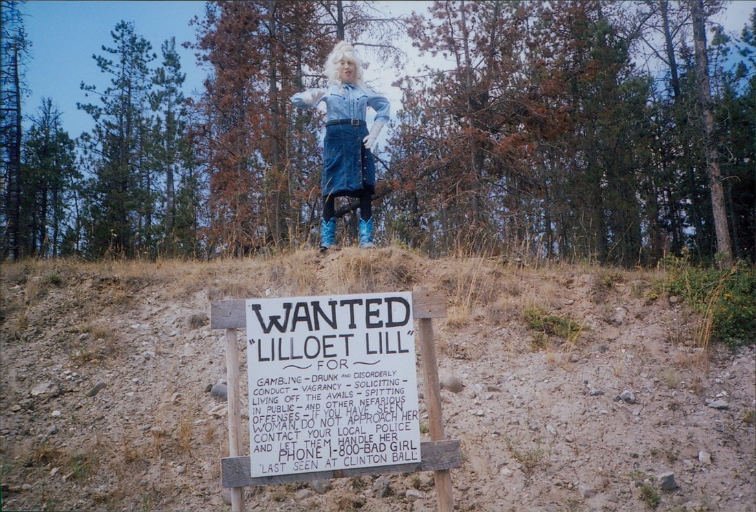 Lillooet Lill in a blond wig, denim western outfit, long gloves and blue cowboy boots, standing on the ridge above her wanted sign