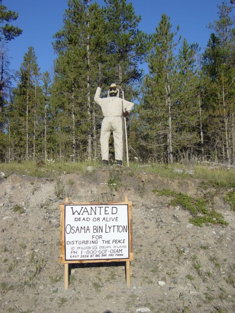 A robed figure with a walking staff stands on the ridge above the Osama Bin Lytton wanted sign, one arm raised in greeting